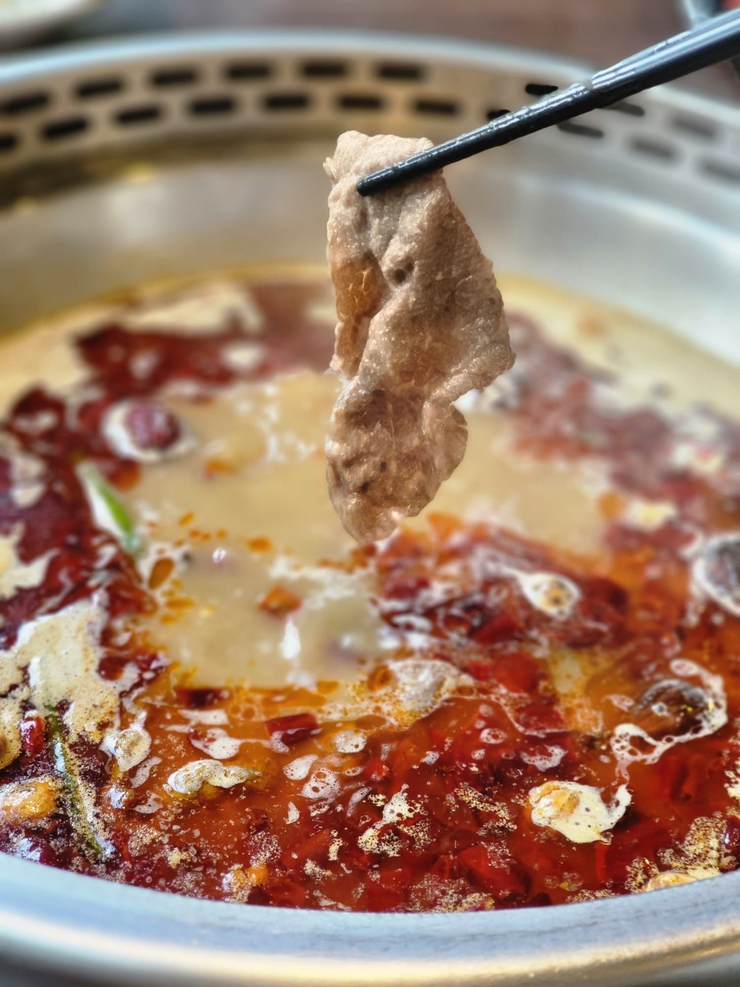 A thin slice of meat lifted with chopsticks from a split pot of spicy broth and bone broth