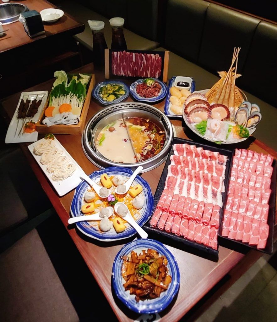A hot pot table seen from above: a split pot surrounded by sliced lamb and beef, seafood, skewers, noodles, vegetables and side dishes