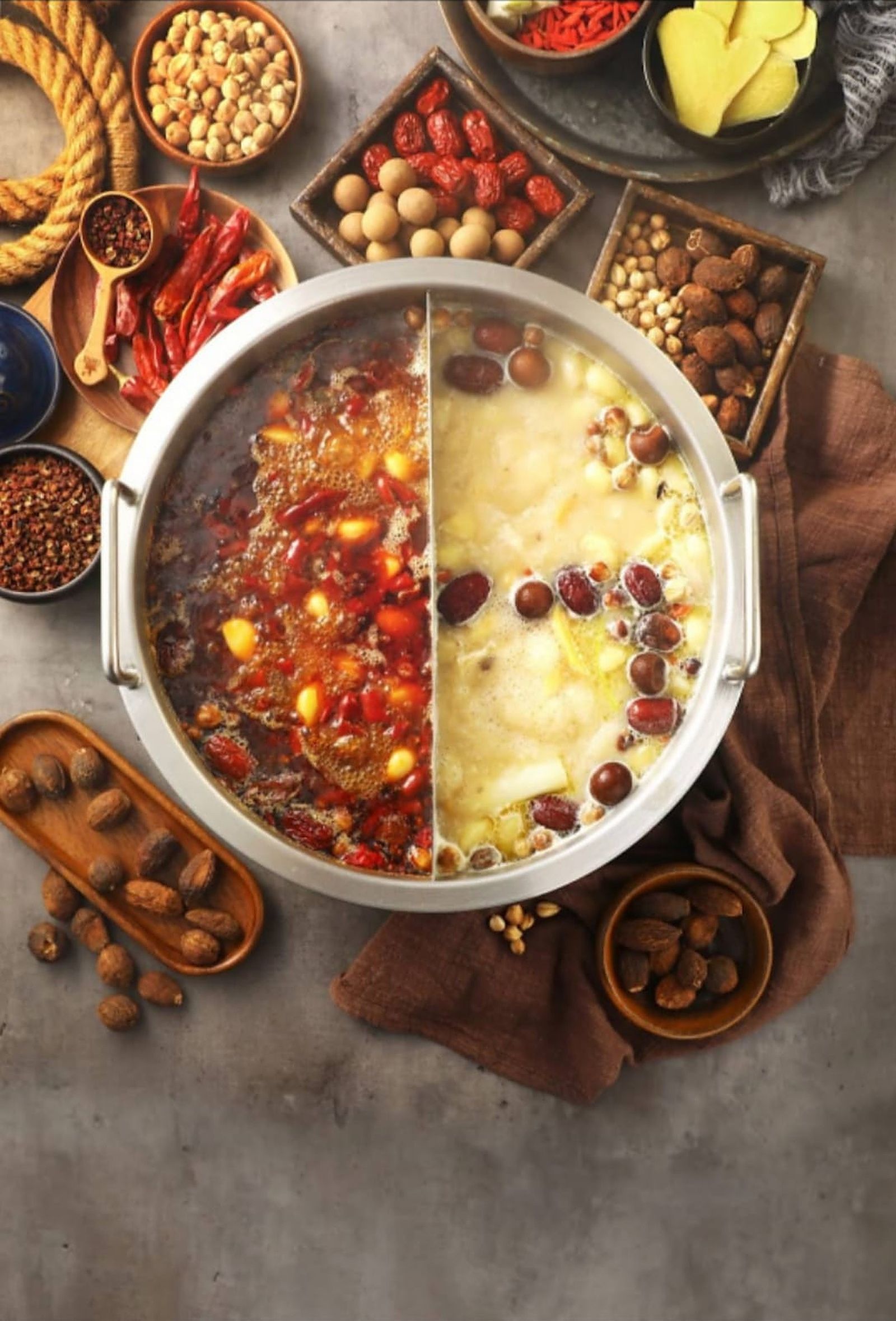 A divided hot pot from above: spicy broth on one side, herbal bone broth with jujubes on the other, surrounded by chillies, goji berries and spices